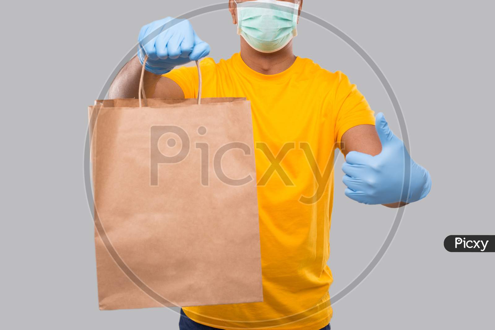Delivery Man With Paper Bag In Hands Wearing Medical Mask And Gloves Showing Thumb Up Isolated Close Up. Yellow Uniform Indian Delivery Boy. Home Food Delivery. Paper Bag