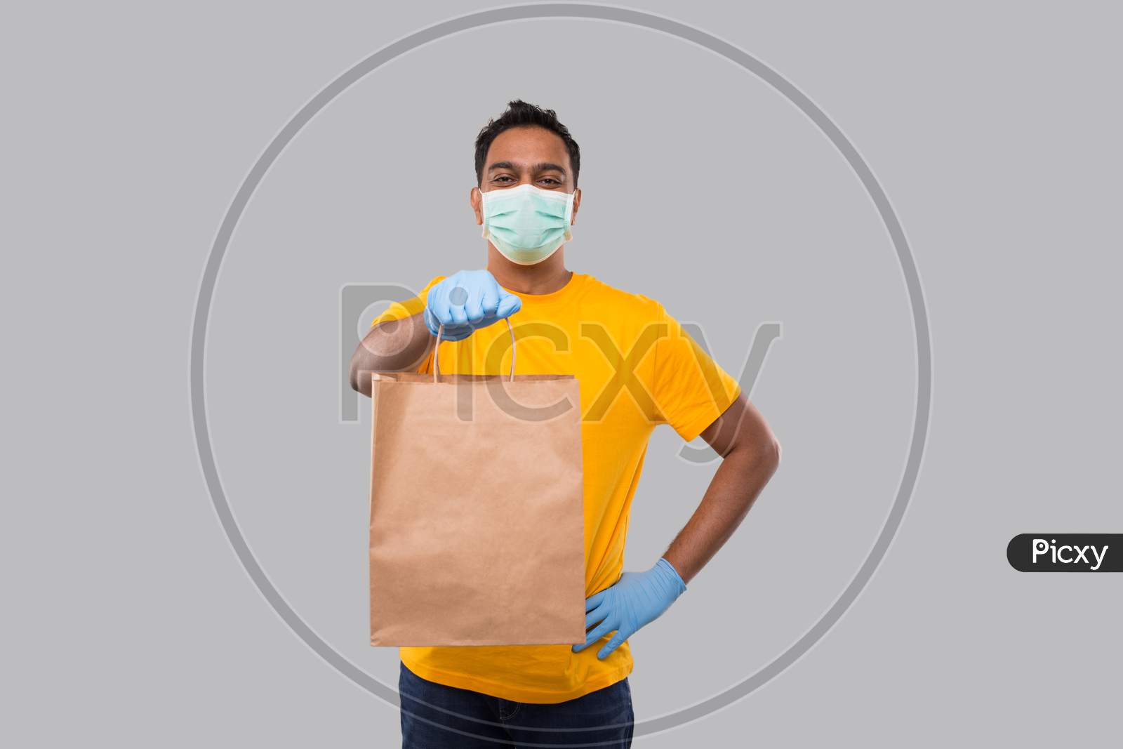 Delivery Man With Paper Bag In Hands Wearing Medical Mask And Gloves Isolated. Yellow Uniform Indian Delivery Boy. Home Food Delivery. Paper Bag