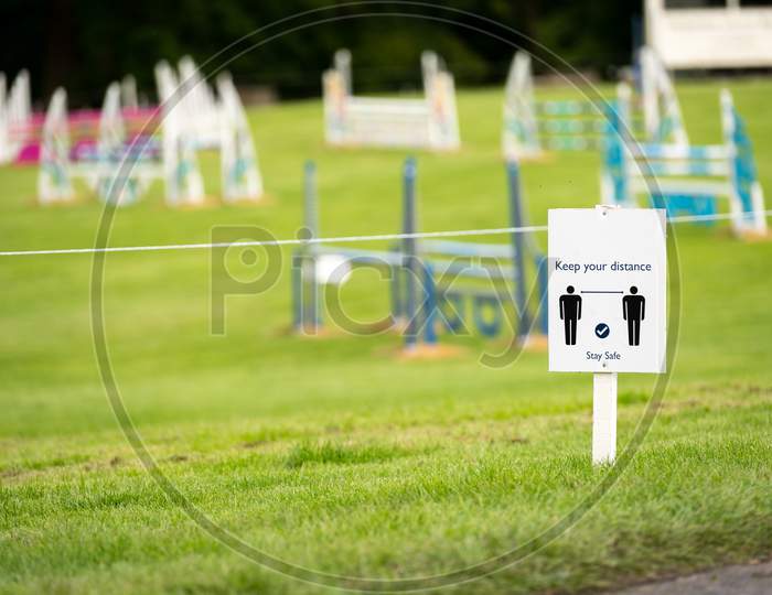 A Covid-19 Social Distancing Sign Staked Into Grass With An Equestrian Show Jumping Outdoor Event In The Background