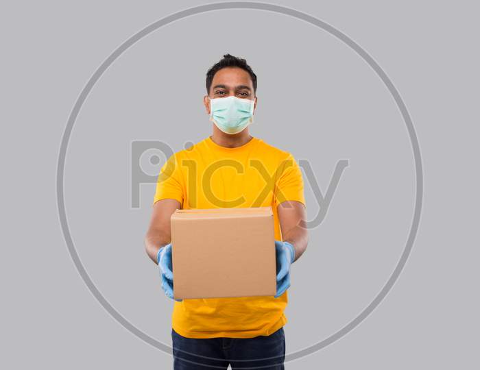 Indian Man With Box In Hands Wearing Medical Mask And Gloves Isolated. Yellow Tshirt Delivery Boy. Home Delivery. Quarantine Hero. Man Smiling
