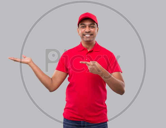 Delivery Man Showing And Pointing To Side Watching In Camera. Indian Delivery Boy In Red Uniform Isolated