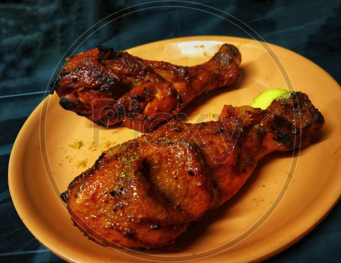 2 Tandoori Chicken Leg Pieces Placed In An Orange Plate With A Piece Of Lemon On A Black Table.