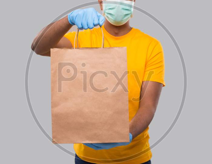 Delivery Man With Paper Bag In Hands Wearing Medical Mask And Gloves Isolated Close Up. Yellow Uniform Indian Delivery Boy. Home Food Delivery. Paper Bag