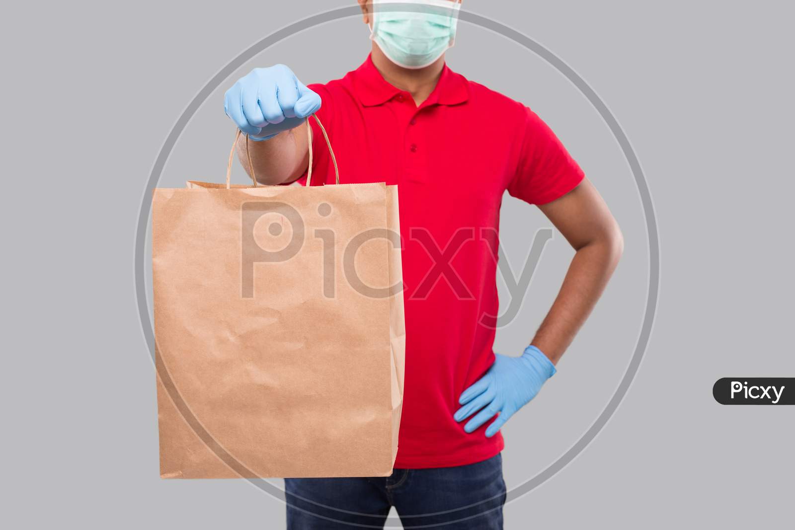 Delivery Man With Paper Bag In Hands Wearing Medical Mask And Gloves Isolated Close Up. Red Uniform Indian Delivery Boy. Home Food Delivery. Paper Bag
