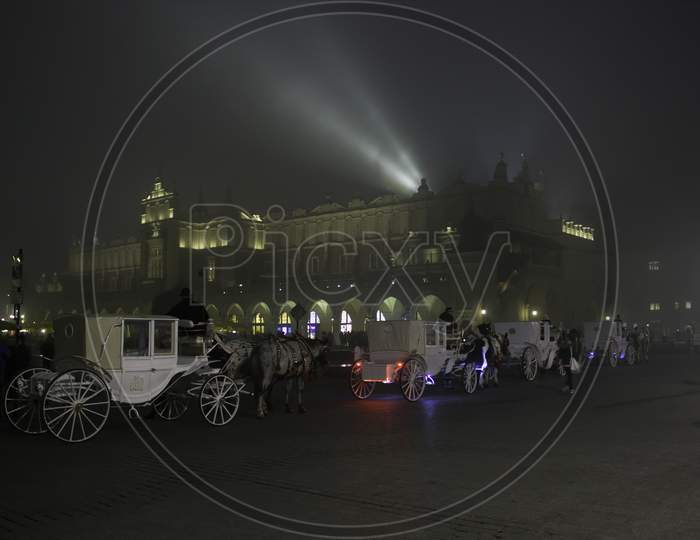 Krakow, Poland - October 29, 2014: Wide Angle View Of Main Square At Center In A Foggy Winter Night With Horse Carts Waiting In A Queue For Tourists