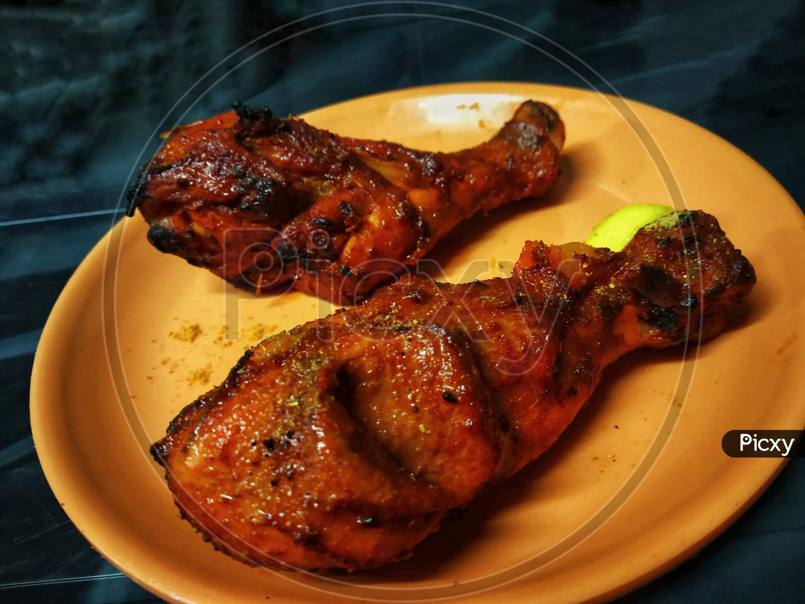 2 Tandoori Chicken Leg Pieces Placed In An Orange Plate With A Piece Of Lemon On A Black Table.