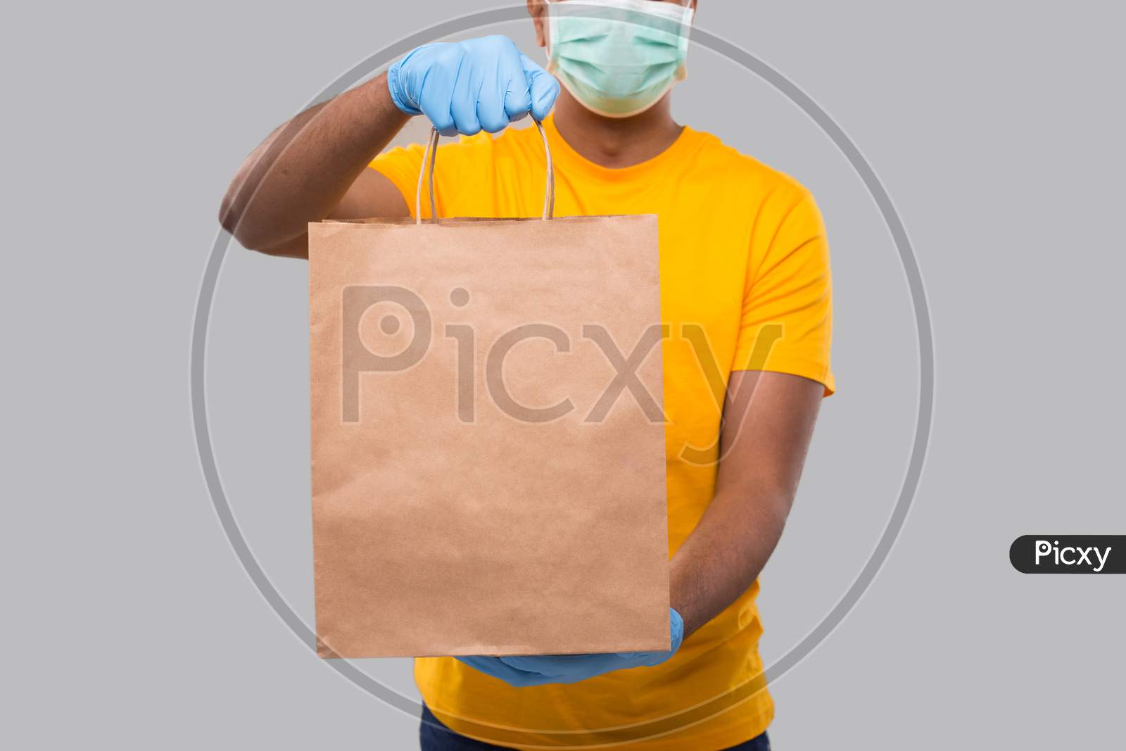 Delivery Man With Paper Bag In Hands Wearing Medical Mask And Gloves Isolated Close Up. Yellow Uniform Indian Delivery Boy. Home Food Delivery. Paper Bag