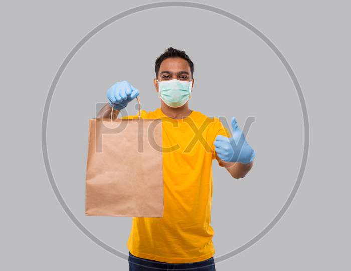 Delivery Man With Paper Bag In Hands Wearing Medical Mask And Gloves Showing Thumb Up Isolated. Yellow Uniform Indian Delivery Boy. Home Food Delivery. Paper Bag