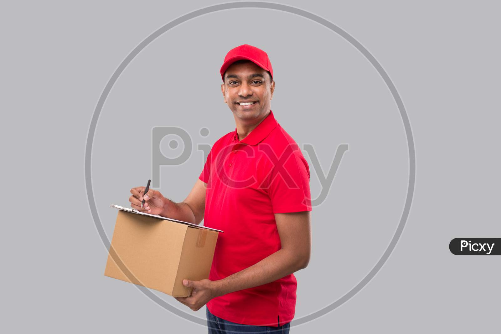 Delivery Man Writing In Clipboard Holding Box Watching In Camera. Indian Delivery Boy Clipboard