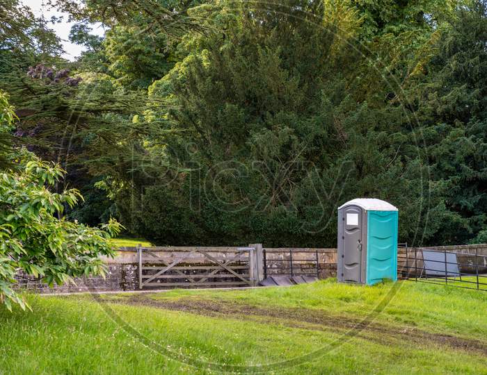 A Plastic Portable Toilet Next To A Muddy Track In A Field At An Outdoor Event And Surrounded By Tall Trees
