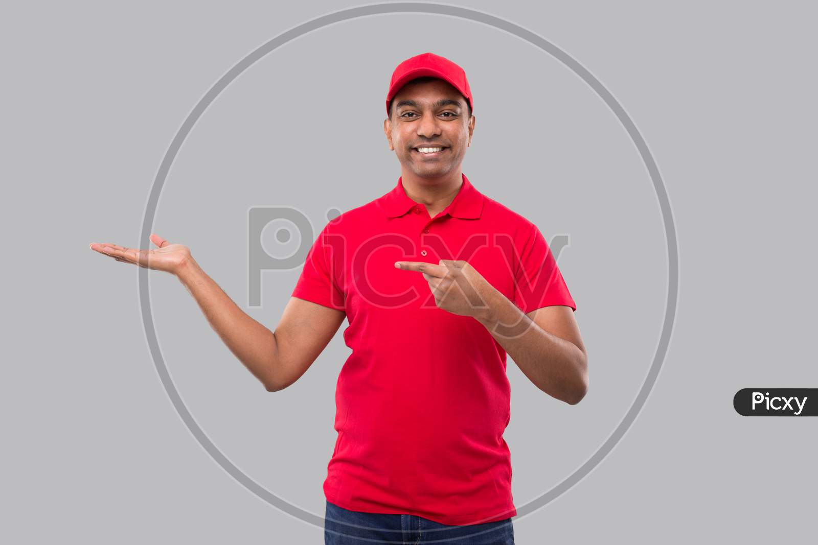Delivery Man Showing And Pointing To Side Watching In Camera. Indian Delivery Boy In Red Uniform Isolated