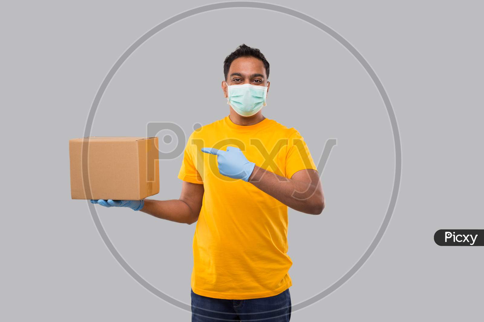 Indian Man Pointing At Box Wearing Medical Mask And Gloves Isolated. Yellow Tshirt Delivery Boy. Home Delivery. Quarantine Hero.
