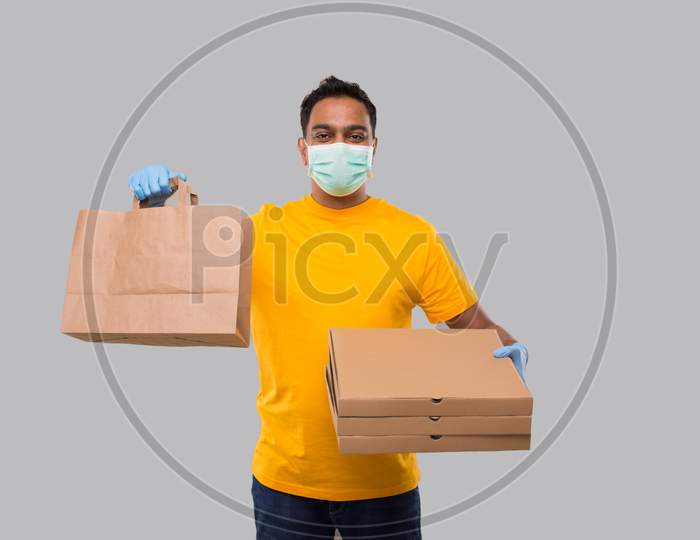 Delivery Man With Paper Bag And Three Pizza Box In Hands Wearing Medical Mask And Gloves Isolated. Yellow Uniform Indian Delivery Boy. Home Food Delivery. Paper Bag