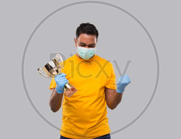 Indian Man Very Happy And Excited, Raising Arms, Celebrating A Victory Or Success Holding Trophy Wearing Medical Mask And Gloves. Winner Sign. Indian Man Isolated With Trophy In Hands