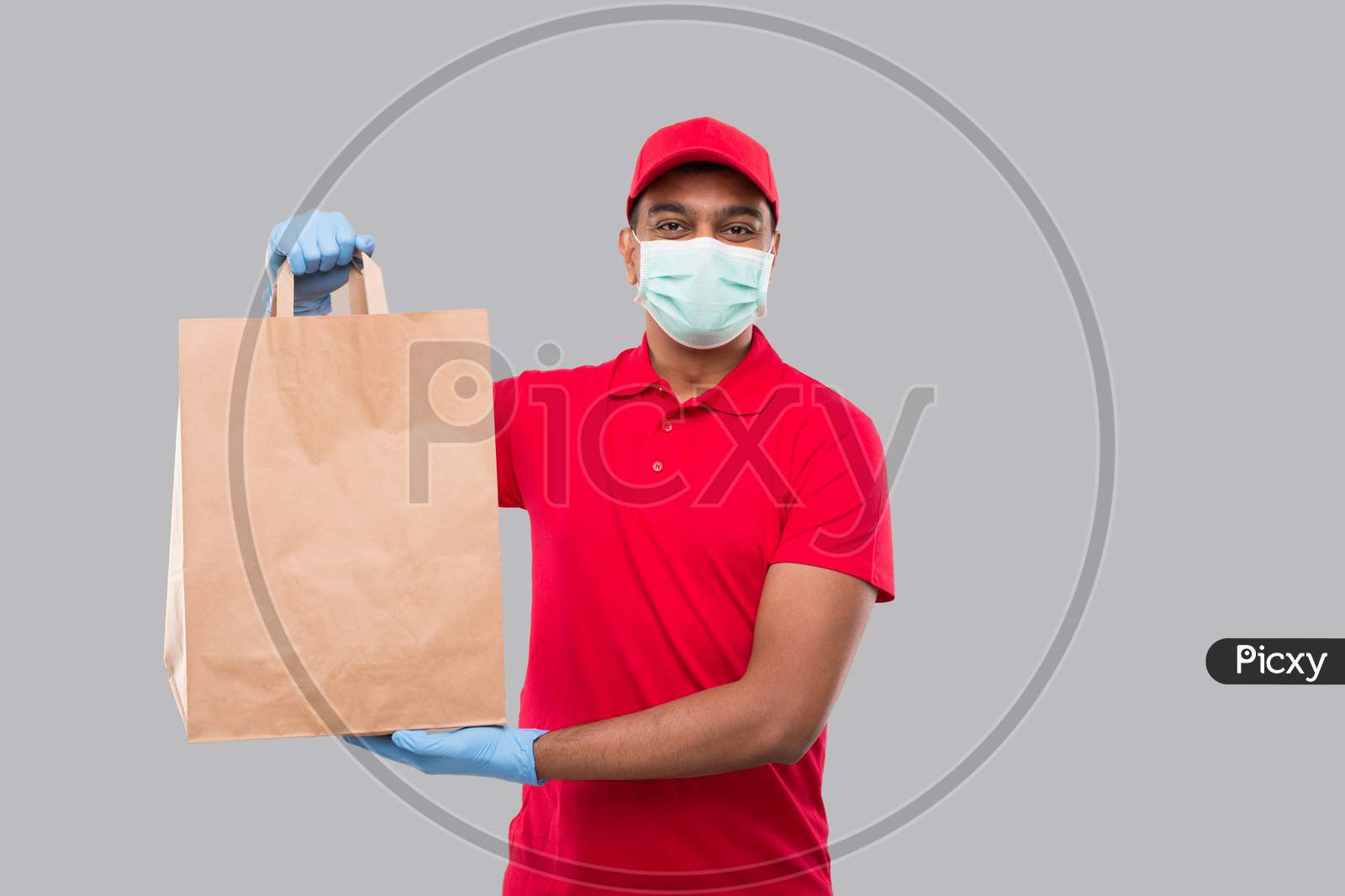 Delivery Man With Paper Bag In Hands Wearing Medical Mask And Gloves Isolated. Red Uniform Indian Delivery Boy. Home Food Delivery. Paper Bag