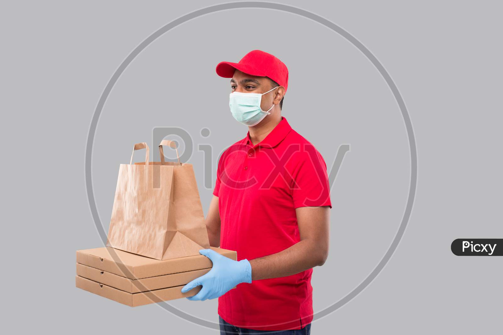 Delivery Man With Paper Bag And Three Pizza Box In Hands Wearing Medical Mask And Gloves Watching Side Isolated. Red Uniform Indian Delivery Boy. Home Food Delivery. Paper Bag