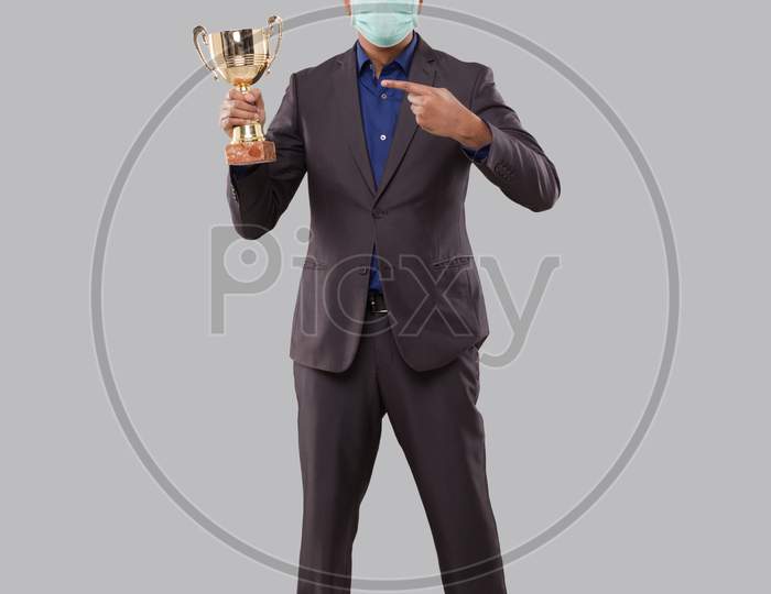 Businessman Pointing At Trophy Wearing Medical Mask. Indian Business Man Standing Full Length With Trophy In Hands