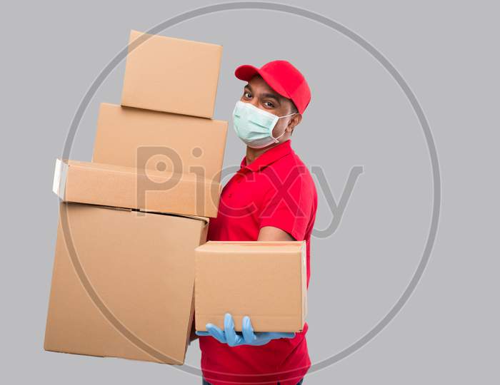 Delivery Man Holding A Lot Carton Boxes Wearing Medical Mask And Gloves Isolated. Indian Delivery Boy Overloaded With Boxes. Gives Out Box To Client