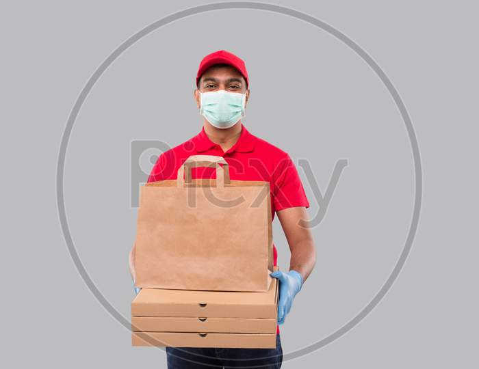 Delivery Man With Paper Bag And Three Pizza Box In Hands Wearing Medical Mask And Gloves Watching In Camera Isolated. Red Uniform Indian Delivery Boy. Home Food Delivery. Paper Bag