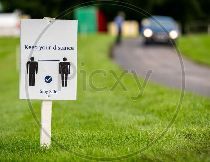 A Covid-19 Social Distancing Sign Staked Into Grass Next To A Road At An Outdoor Event