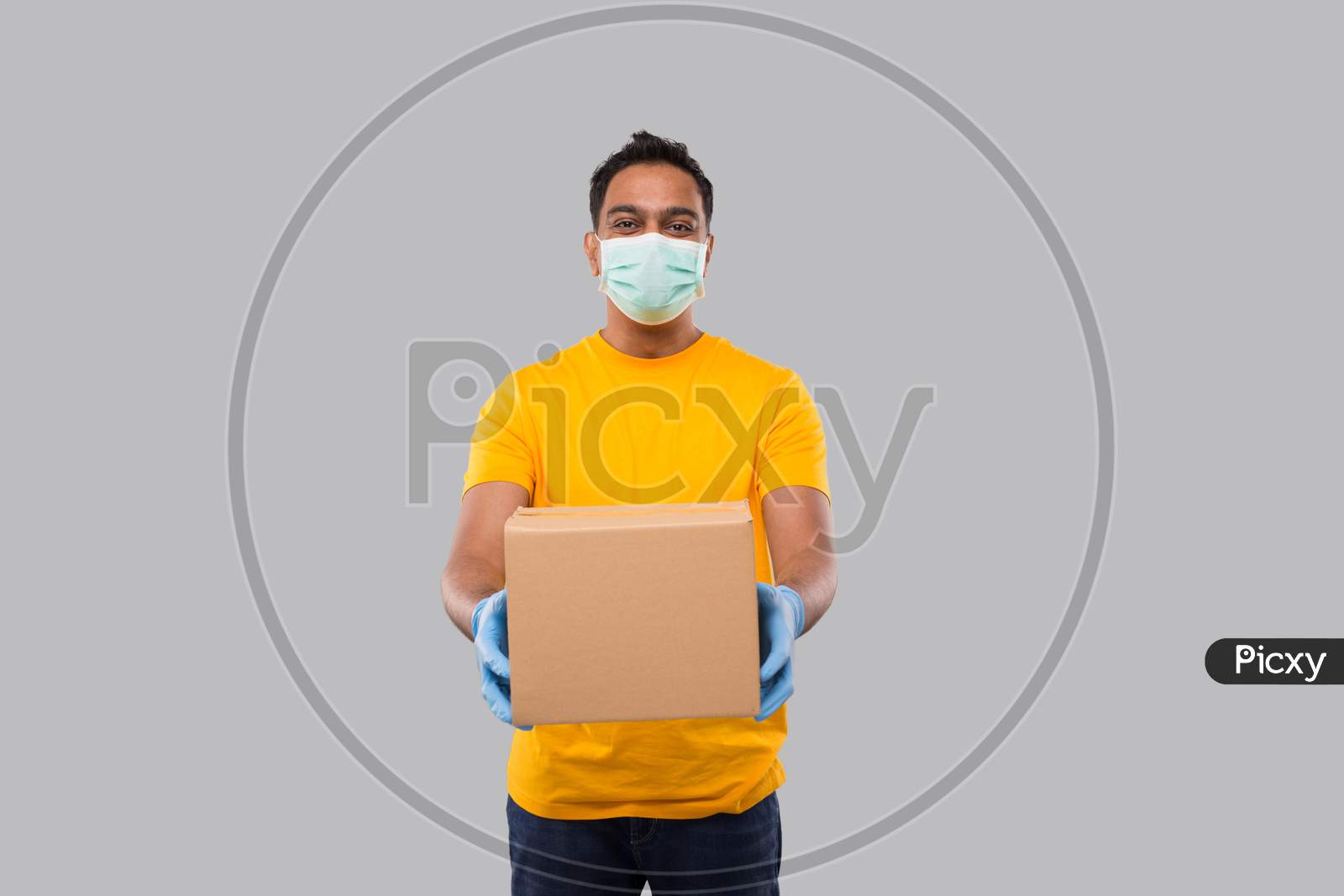 Indian Man With Box In Hands Wearing Medical Mask And Gloves Isolated. Yellow Tshirt Delivery Boy. Home Delivery. Quarantine Hero. Man Smiling