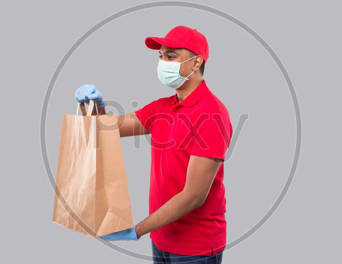 Delivery Man With Paper Bag In Hands Wearing Medical Mask And Gloves Watching Side Isolated. Red Uniform Indian Delivery Boy. Home Food Delivery. Paper Bag