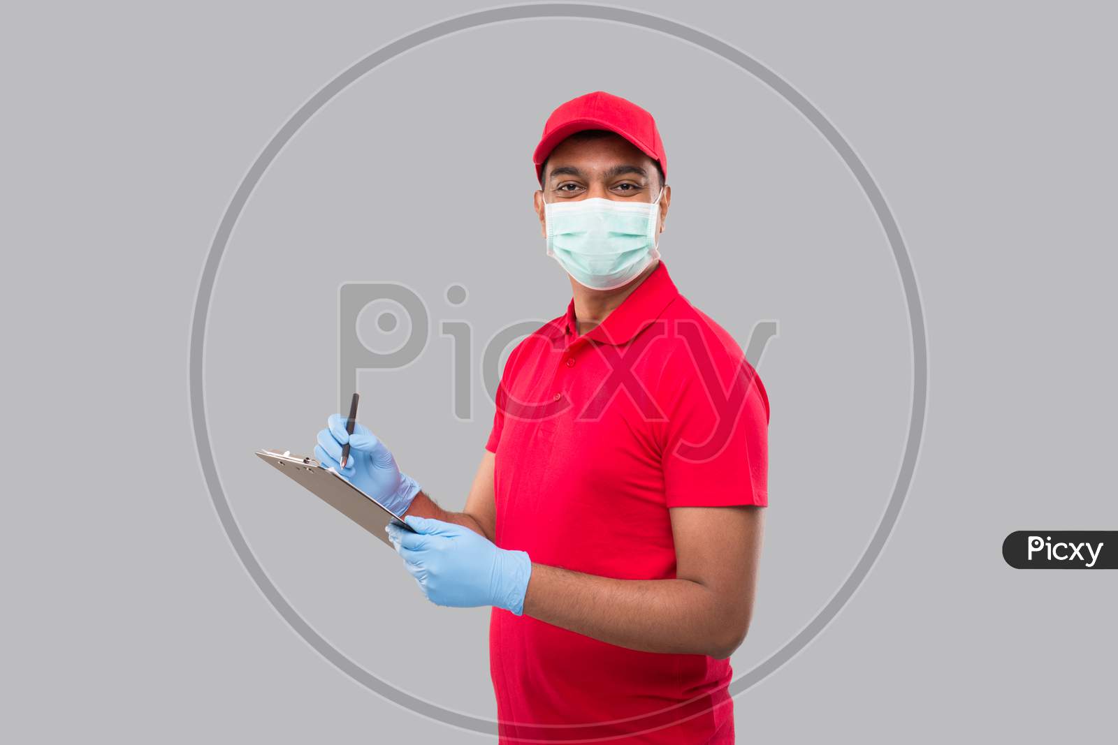 Delivery Man Writing In Clipboard Wearing Medical Mask And Gloves Watching In Camera. Indian Delivery Boy Clipboard
