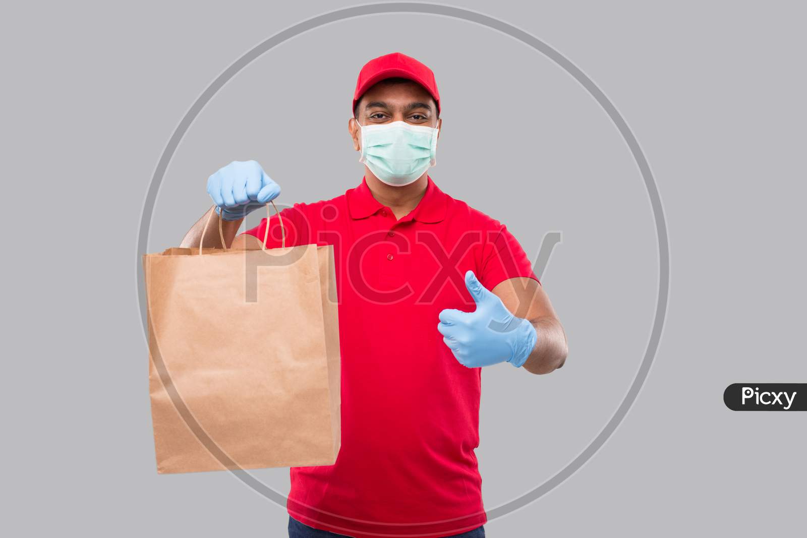 Delivery Man With Paper Bag In Hands Wearing Medical Mask And Gloves Showing Thumb Up Isolated. Red Uniform Indian Delivery Boy. Home Food Delivery. Paper Bag
