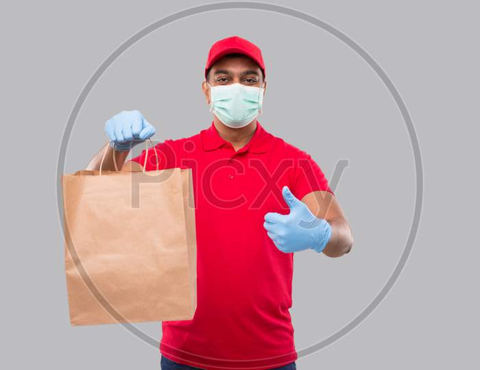 Delivery Man With Paper Bag In Hands Wearing Medical Mask And Gloves Showing Thumb Up Isolated. Red Uniform Indian Delivery Boy. Home Food Delivery. Paper Bag