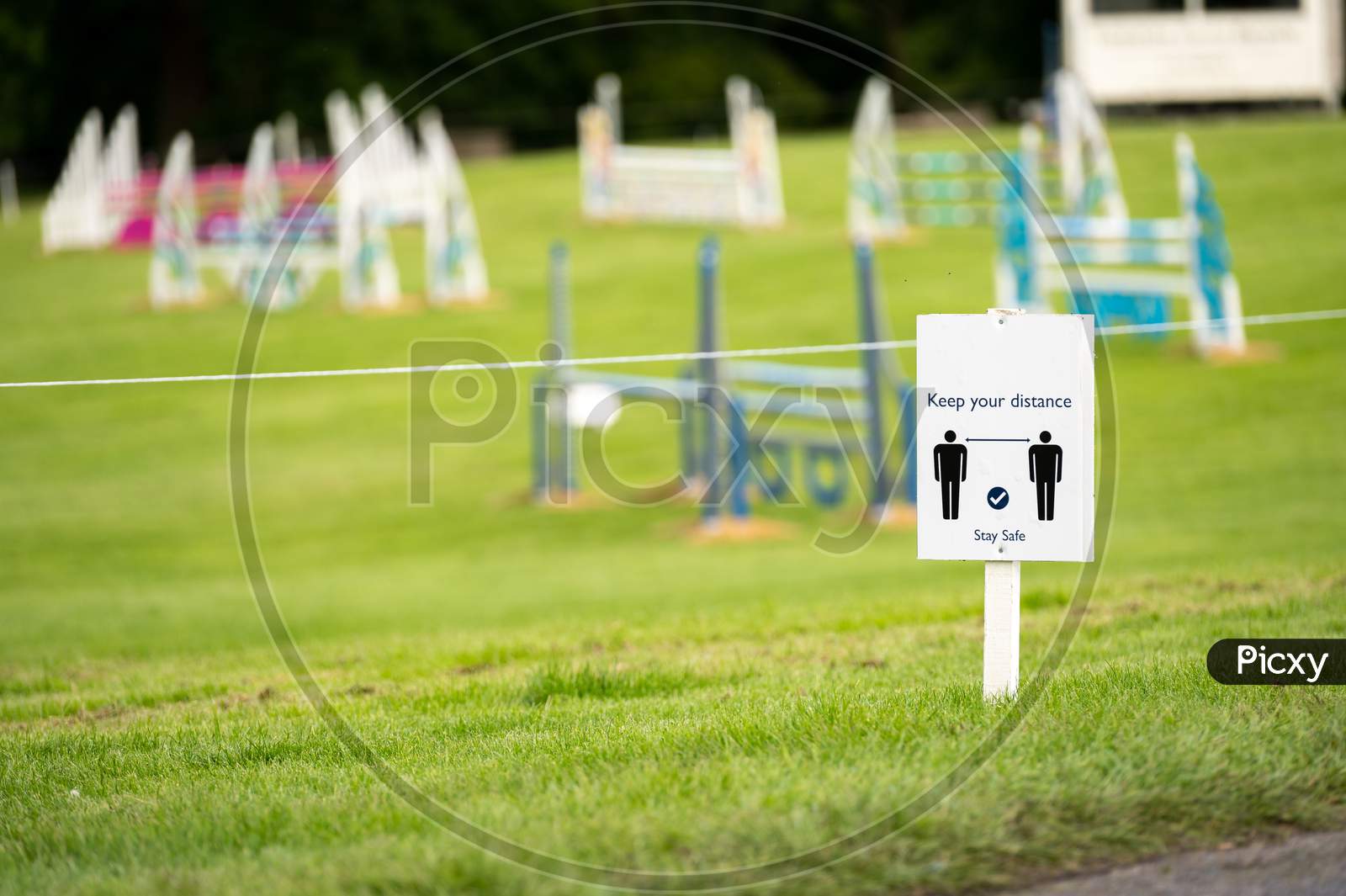 A Covid-19 Social Distancing Sign Staked Into Grass With An Equestrian Show Jumping Outdoor Event In The Background