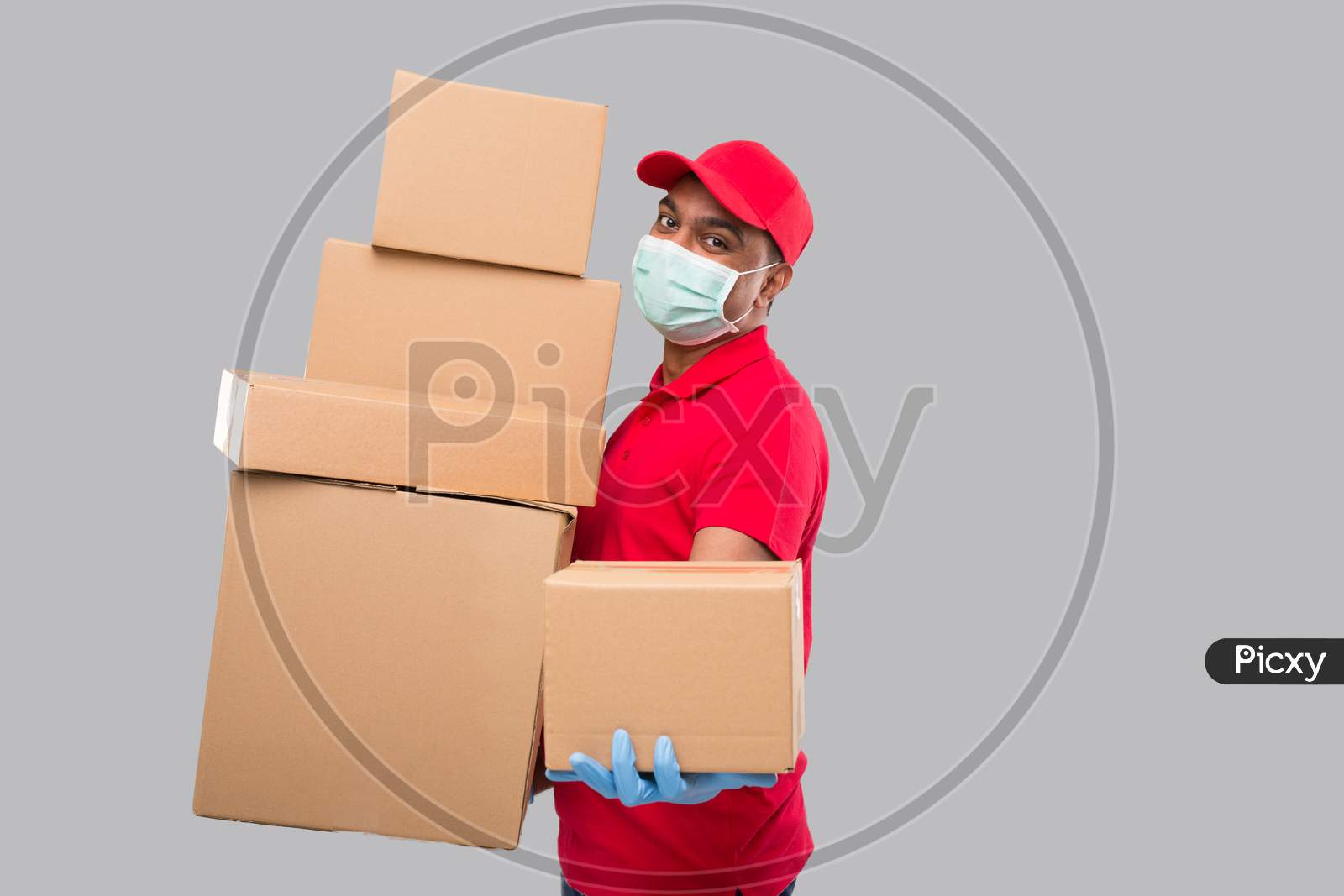 Delivery Man Holding A Lot Carton Boxes Wearing Medical Mask And Gloves Isolated. Indian Delivery Boy Overloaded With Boxes. Gives Out Box To Client