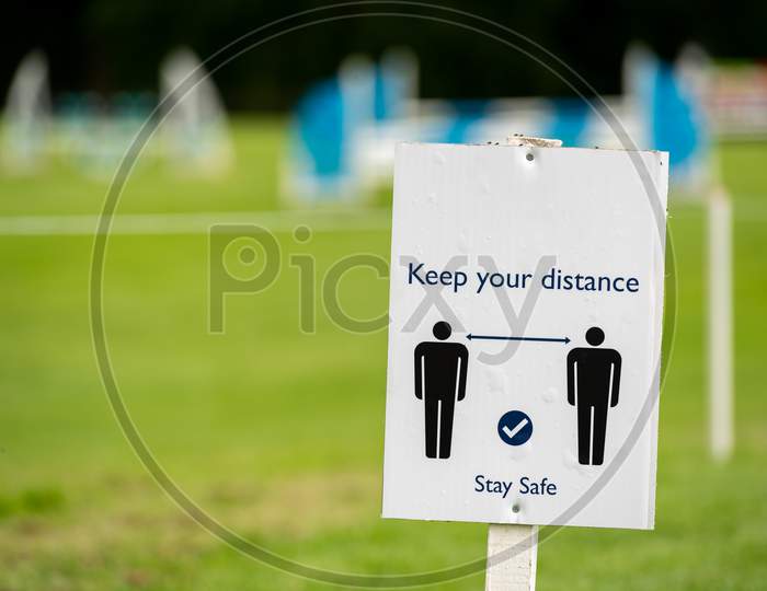 A Covid-19 Social Distancing Sign With An Out Of Focus Outdoor Event In The Background