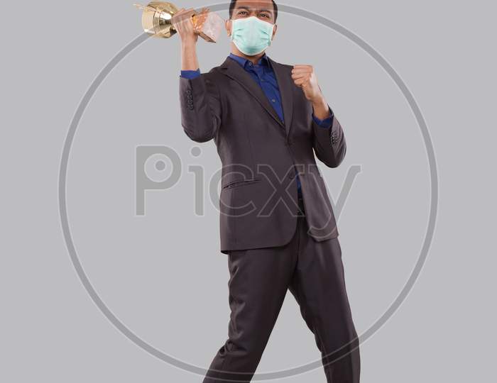 Businessman Very Happy And Excited, Raising Arms, Celebrating A Victory Or Success Holding Trophy Wearing Medical Mask. Winner Sign. Indian Business Man Isolated With Trophy In Hands