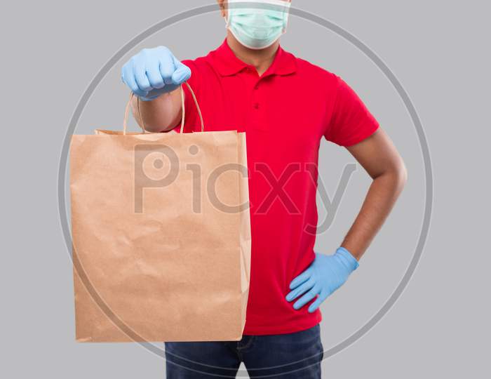 Delivery Man With Paper Bag In Hands Wearing Medical Mask And Gloves Isolated Close Up. Red Uniform Indian Delivery Boy. Home Food Delivery. Paper Bag