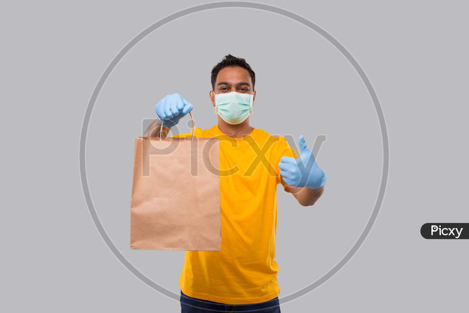 Delivery Man With Paper Bag In Hands Wearing Medical Mask And Gloves Showing Thumb Up Isolated. Yellow Uniform Indian Delivery Boy. Home Food Delivery. Paper Bag