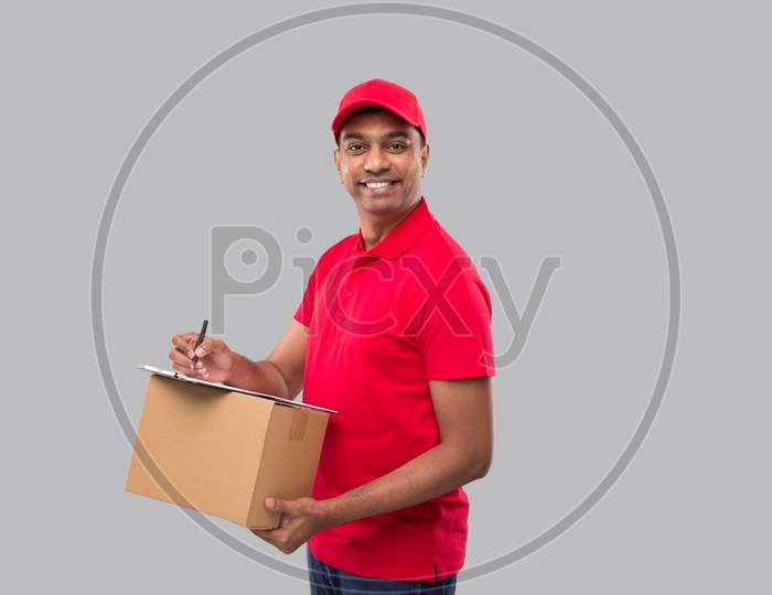 Delivery Man Writing In Clipboard Holding Box Watching In Camera. Indian Delivery Boy Clipboard