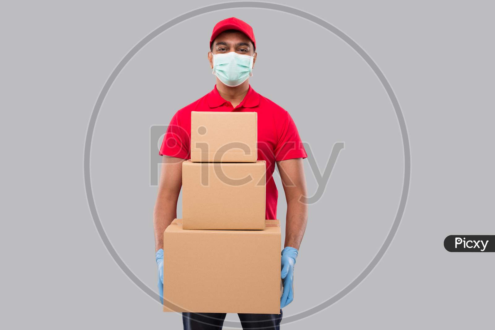 Delivery Man Holding Carton Boxes Wearing Medical Mask And Gloves Isolated. Indian Delivery Boy Smiling With Boxes In Hands