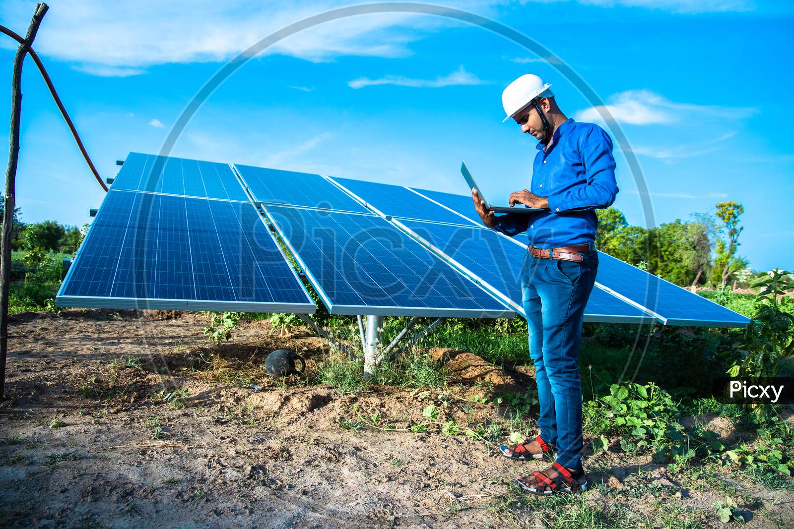 Young Male Engineer With Laptop In Hand Standing Near Solar Panels, Agriculture Farm Land With Clear Blue Sky Background, Renewable Energy, Clean Energy.