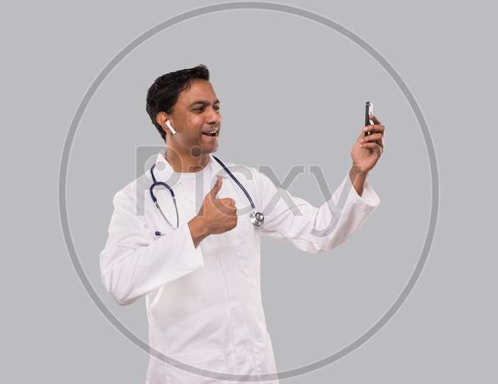 Doctor Having Video Call On Phone Showing Thumb Up With Wireless Earphone Isolated. Indian Man Doctor Video Call. Medicine Online. Doctor Using Phone.