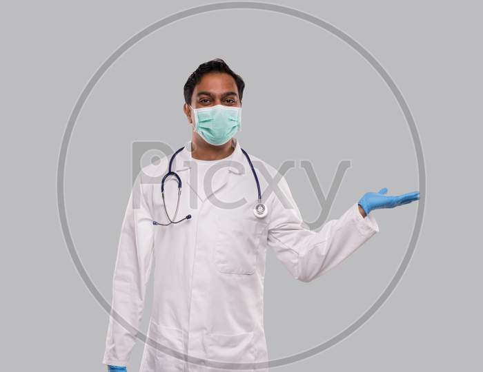 Doctor Holding Hand To Side Wearing Medical Mask And Gloves Watching To Camera Isolated. Indian Man Doctor Sign
