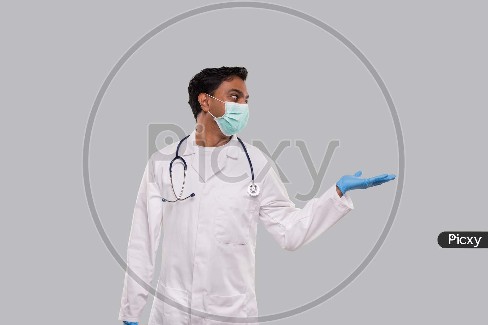 Doctor Holding Hand To Side Wearing Medical Mask And Gloves Watching To Side Isolated. Indian Man Doctor Sign