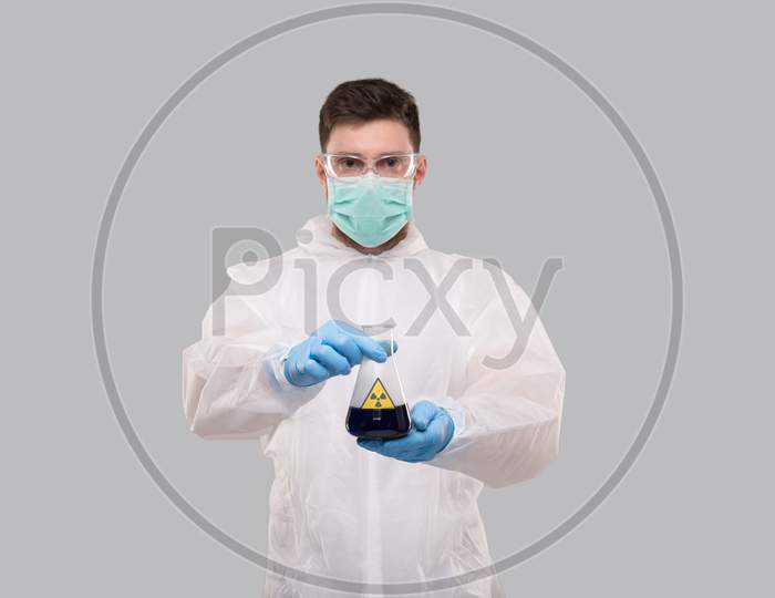 Male Laboratory Worker In Chemical Suit, Wearing Medical Mask And Glasses Showing Flask With Blue Liquid Radiation Sign. Science, Medical, Virus Concept