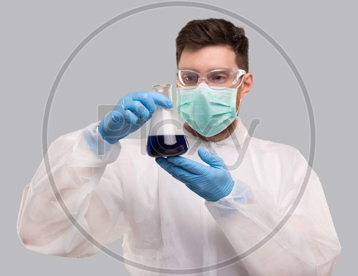 Male Laboratory Worker In Chemical Suit, Wearing Medical Mask And Glasses Watching Flask With Colorfull Liquid. Science, Medical, Virus Concept. Blue Liquid