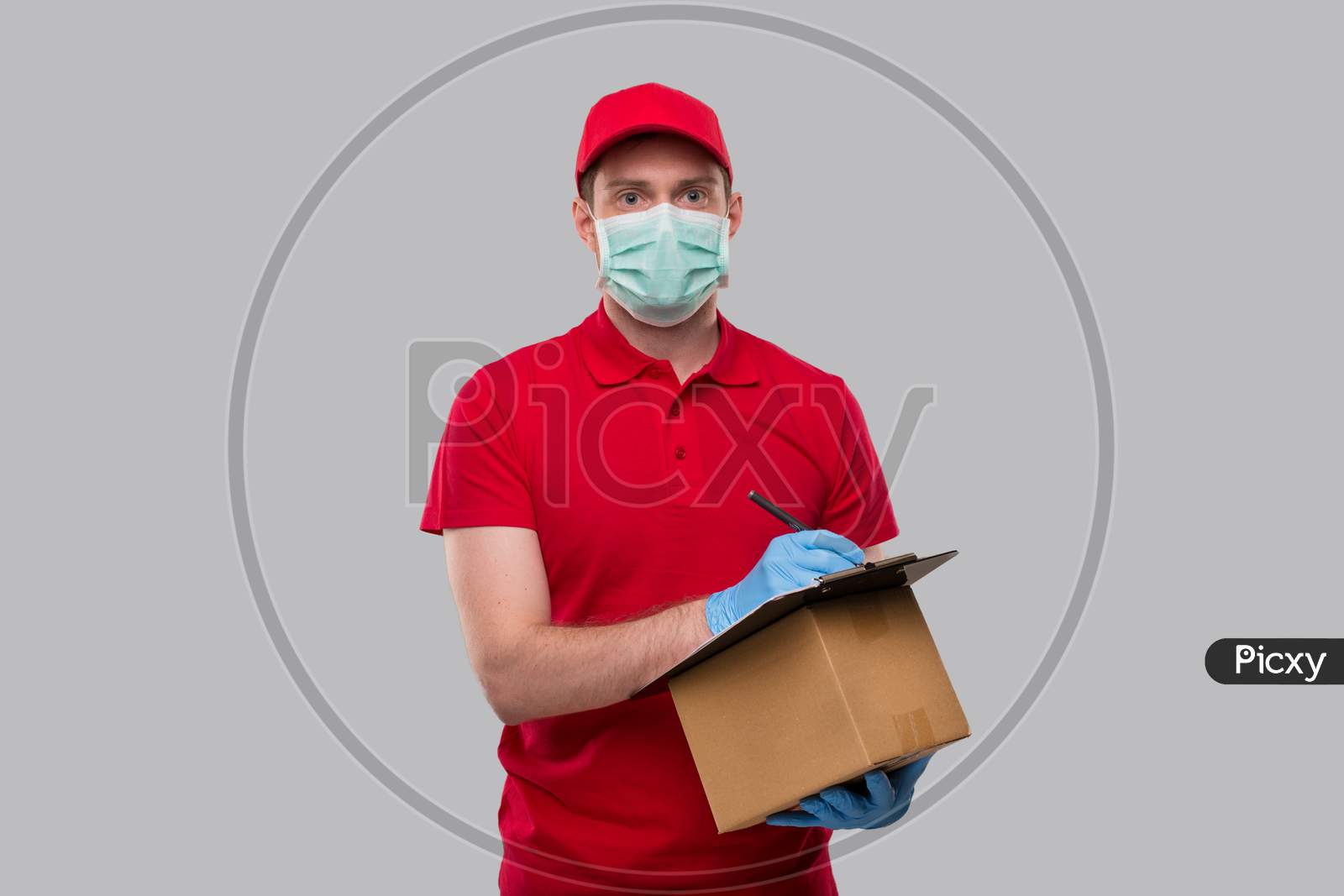 Delivery Man Wearing Medical Mask And Gloves Writing In Clipboard With Box In Hands Watching In Camera. Delivery Boy Home Delivery.