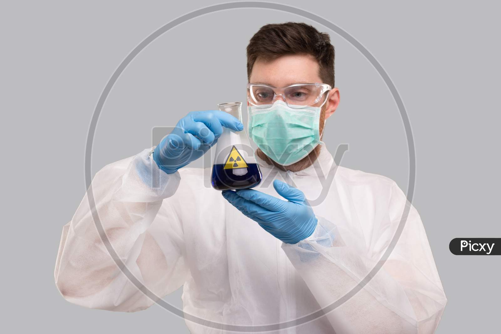 Male Laboratory Worker In Chemical Suit, Wearing Medical Mask And Glasses Watching Flask With Blue Liquid Radiation Sign. Science, Medical, Virus Concept. Blue Liquid