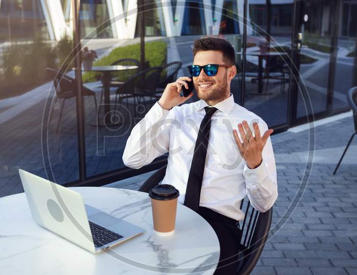 Businessman Working In Cafe With Laptop Drinking Coffee On A Sunny Day. Business Man Talking On Phone Smilling On Business Lunch.