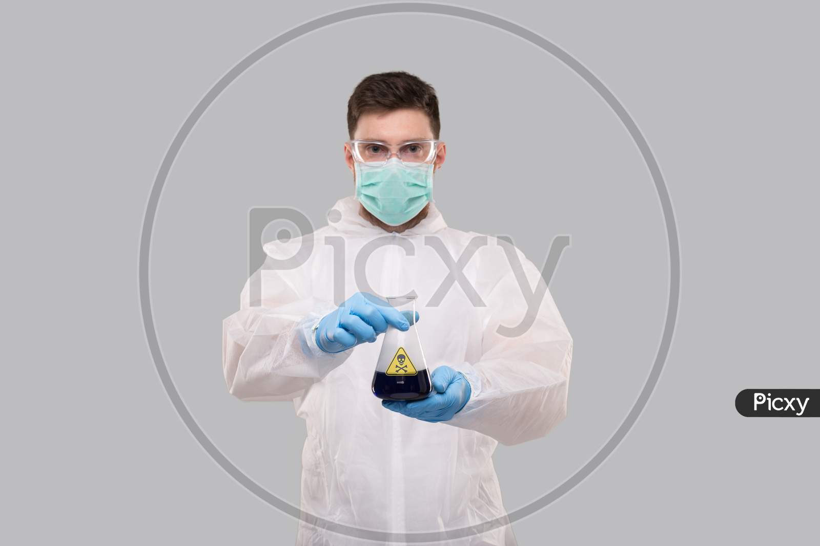 Male Laboratory Worker In Chemical Suit, Wearing Medical Mask And Glasses Showing Flask With Blue Liquid Poison Sign. Science, Medical, Virus Concept