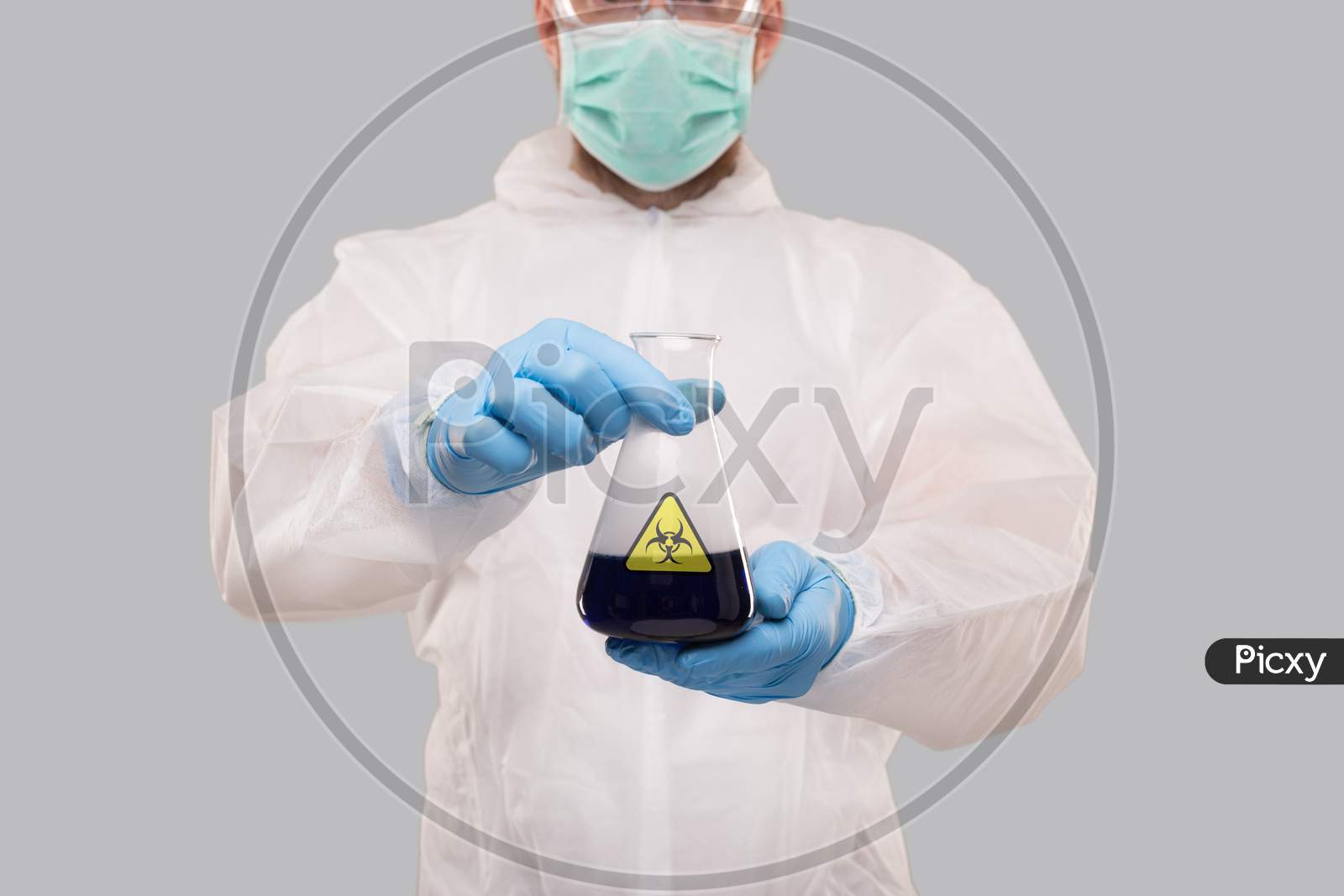 Male Laboratory Worker In Chemical Suit, Wearing Medical Mask And Glasses Showing Flask With Blue Liquid Biohazard Sign. Science, Medical, Virus Concept