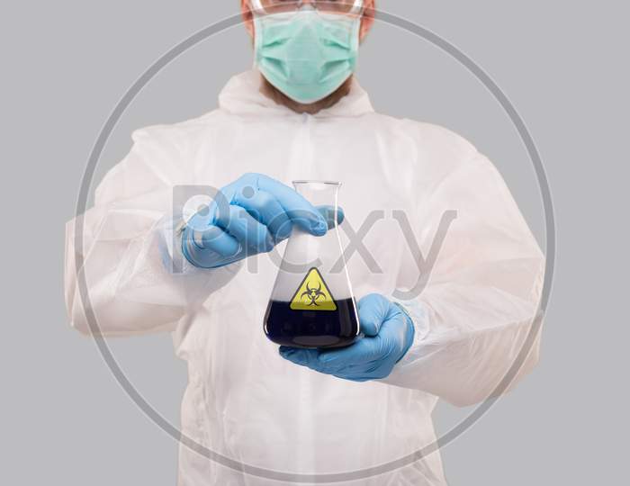 Male Laboratory Worker In Chemical Suit, Wearing Medical Mask And Glasses Showing Flask With Blue Liquid Biohazard Sign. Science, Medical, Virus Concept
