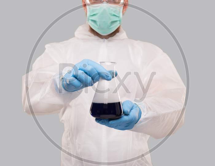 Male Laboratory Worker In Chemical Suit, Wearing Medical Mask And Glasses Showing Flask With Colorfull Liquid. Science, Medical, Virus Concept